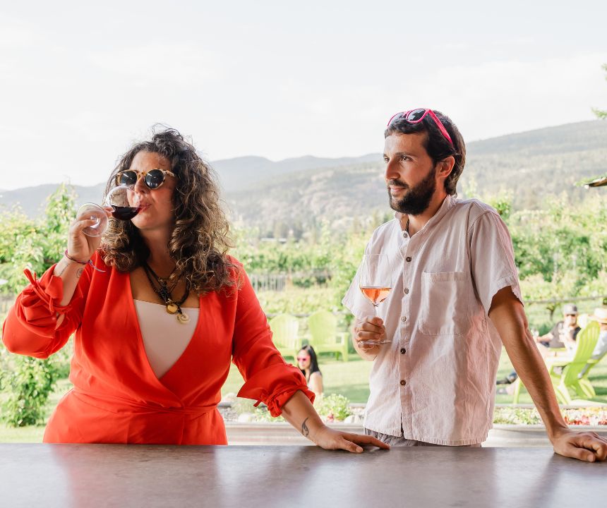 A women in a red dress sipping red wine with a gentleman wearing JoieFarm sunnies and holding a glass of blush wine outdoors at JoieFarm