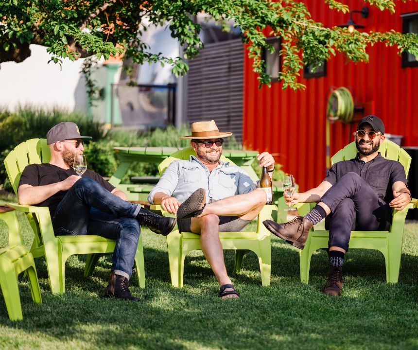 3 smiling gentlemen sitting on green patio chairs on JoieFarm's Picnique lawn with glasses of JoieFarm wine