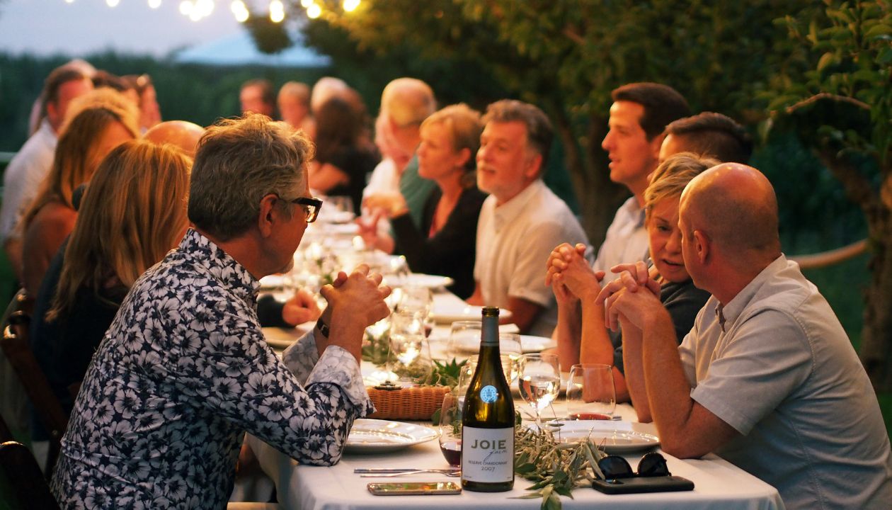 Group of people sitting at an evening lit long table enjoying each other and JoieFarm wine.